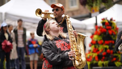 A young girl with blonde hair plays a trumpet in a city square. 