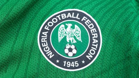 A close up of the Nigeria Football Federation's circular crest sitting on a green shirt. The crest depicts a white eagle perched on a black and white football on a green circular background which is surrounded by the wording Nigeria Football Federation 1945 written in white text on a black background which goes around the outside of the design