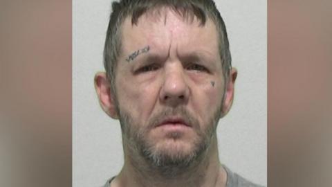 Mugshot of McKenna. He has a lined face, untidy short brown hair, a grey beard and tattooed symbols above his right eye and a heart tattooed on his left cheek.