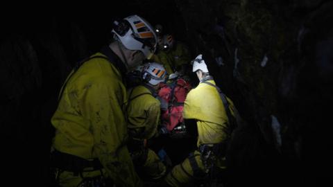 Underground rescue team COMRU in yellow overalls surround a red-blanketed stretcher inside a cave