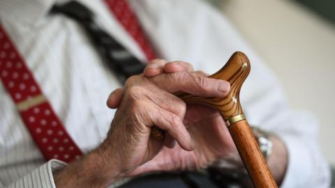 Generic image which shows a close-up of an elderly man's hands clasping the top of his walking stick. He is sitting down and wears a white striped shirt, red pattenered braces and dark tie.