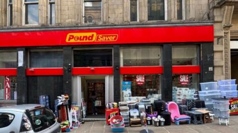 A stone Victorian building front with red signage and yellow Pound Saver lettering. A variety of plastic goods are stacked in front of the shop on the pavement.







