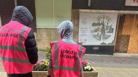 Two boys standing with their backs to the camera, wearing pink high-visibility jackets with the words 'Longton Exchange' written on them. There is a raised flower bed in front of them in a wooden container.