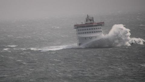 The Manxman ferry in stormy conditions. The white and red vessel is in choppy seas and a large white wave crashes in front of it.