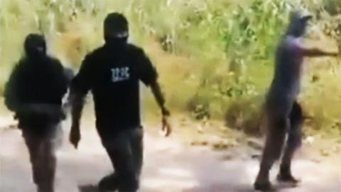 A grainy still of footage featuring two men dressed in black and wearing balaclavas which obscure all but their eyes. They are walking along a dirt path with greenery alongside. To the right is another man wearing a baseball cap