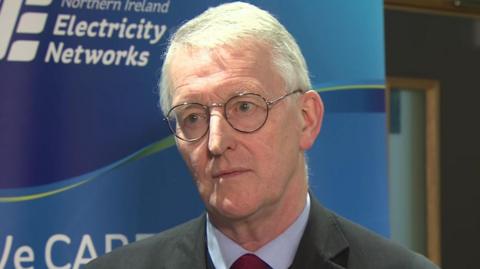 Hillary Benn - a man with white hair and circular glasses. He is standing in front of a blue banner with NI Electricity Networks on it. He is wearing a grey jacket, a blue shirt and a red tie.