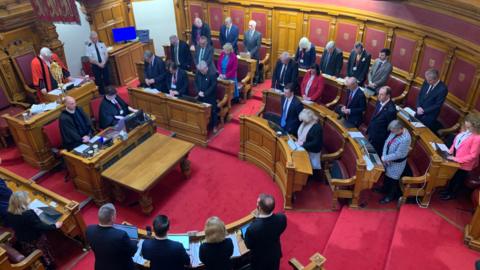 The States Assembly with smartly dressed men and women standing around the chamber with heads bowed. There are wooden panelled walls and desks and a bright red carpet