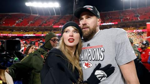 Taylor Swift and Travis Kelce embrace in a stadium. Both wear hats, Travis wears a grey T-shirt with 'Champions' on it