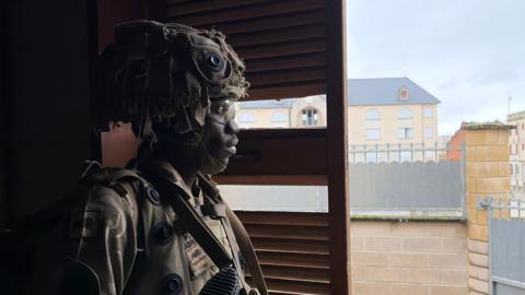 A soldier in helmet and army gear peers out from behind a door shutter to other buildings outside.
