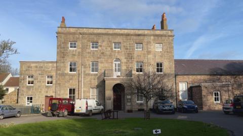 A large, three-storey granite government building with cars parked in front and a grass lawn.