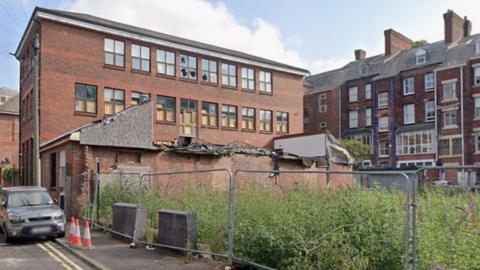 Lanson House, on Mount Street in Preston, is a large three-storey rectangular redbrick building with many broken windows next to a fenced-off area with overgrown weeds.