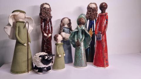 Nativity models made of clay. On the left is a Shepard, wearing green clothes and holding a Shepards crook. To the right of the Shepard is a small sheep. To the right is model of Joseph, wearing brown clothes and with brown hair and a brown beard. To the right is a smaller model of a girl wearing a green dress and with brown hair. To the right is a model of Mary, with brown hair, and holding a baby. To the right are three wise men, one in blue and brown, one in blue and one in red. They are all holding gifts. None of the models have faces.