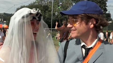 Woman on the left wearing veil and bride tiara, man on the right wearing tie and suit and navy cap