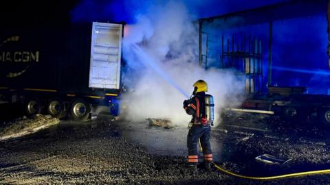 The back of a lorry with a lot of smoke and a firefighter trying to put out the fire.