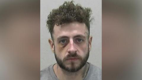 Mugshot of Coe. He has a short dark beard and short thick curly dark hair. He appears to have a bruise around his right eye and an illegible black symbol tattooed beside his left eye