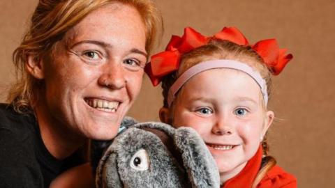 A woman and young child smile at the camera, There is a stuffed grey toy in the middle of them. The child has a pink band around her forehead and is wearing a red top, She has matching hair bobbles.