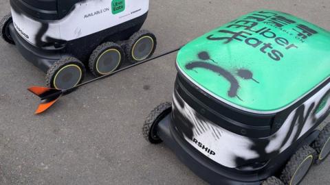 Robotic delivery vehicles with spray paint on them and a visibility flagpole bent to the ground. A sad face has been spray-painted on one of the robots in black paint.
