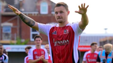 Jayden Wareham is standing with his arms out stretched facing the Exeter City fans. He is wearing the red and white home kit of Exeter.