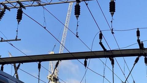 Overhead rail wires against a blue sky. A white crane is in the background