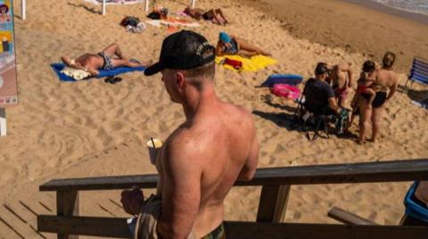 A man with a black cap and no shirt on walks down to a beach in Albufeira, where other people are lying on the yellow sand.