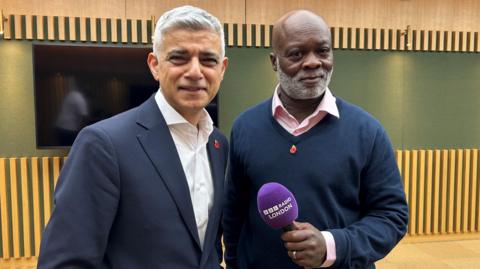 Sadiq Khan in a white shirt and navy suit. he stands next to Eddie Nestor, who is in a pink shirt and navy v-neck jumper. Eddie Nestor holds a 91ȱ London microphone.