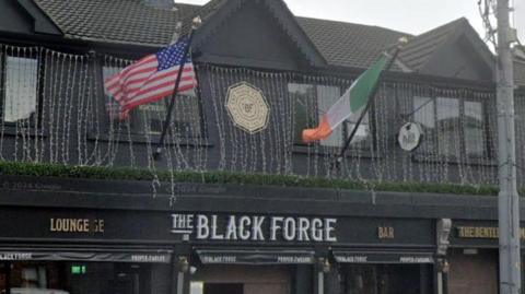 The exterior of The Black Forge pub. The building is painted black with the Black Forge written in white across the entrance. There is an Irish and a US flag flying above the entrance. 