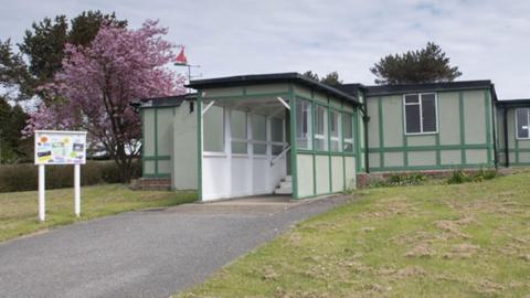 A single-level white building with a black roof and green wooden beams. The sky is overcast and the centre is surrounded by grass.
