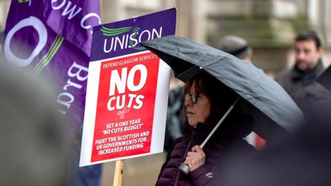 A woman under an umbrella holding a red placard that says No Cuts