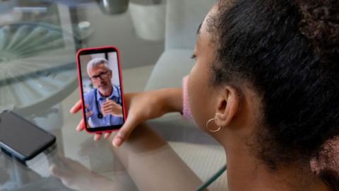 A woman, seen from behind, holds a phone and speaks to it. A doctor wearing a stethoscope round his neck is on the screen