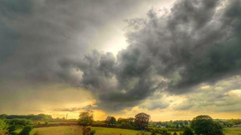 The image shows a rural landscape of fields, hedgerows and scattered trees beneath an expansive sky dominated by dramatic dark-grey storm clouds, with a bright break in the cloud cover near the horizon allowing golden sunlight to spill across the scene.