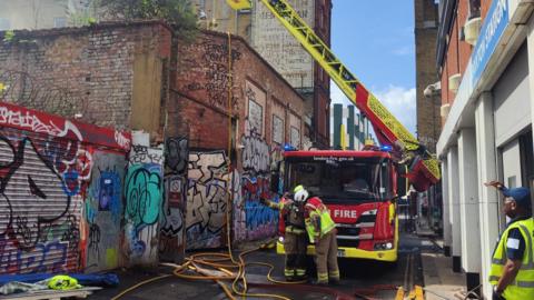 A fire truck and firefighters stand by hoses in a back street in London. There is graffiti on the walls on one side of the road. 