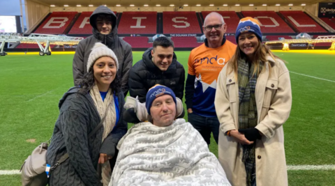 A man in a blue beanie hat siting on a wheelchair with a white blanket on him, surrounded by three men and two women in coats, standing on the pitch at Ashton Gate football stadium