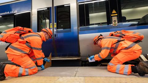 Two men in high-viz work gear kneel next to a train, measuring how far it is from the platform