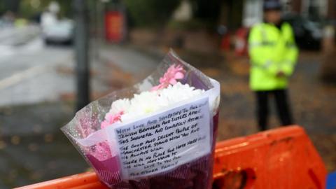A heartfelt tribute placed on an orange barricade. A bouquet of white and pink flowers is wrapped in purple and white paper. Attached to the bouquet is a handwritten note expressing solidarity and sympathy:

"With love and sympathy to our Jewish brothers and sisters after this shameful and cowardly attack. The good and decent people of Manchester stand with you and we always will. There is no place for hate here. We will not have it.
— The O'Shea Family

In the background, a person in a high-visibility jacket stands near the barricade.