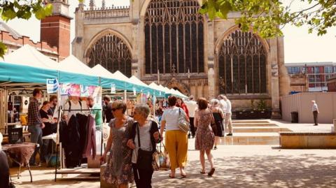 People walking past a row of market stalls in front of large church. The stalls have green canopies and the people are dressed in summer attire. 