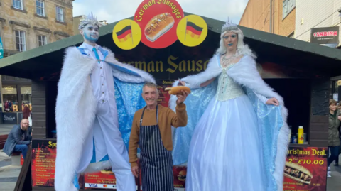 Street artists pose with Charlie Pearson in front of his wooden sausage cabin