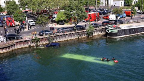 The River Seine in Juvisy-sur-Orge. The roof of a bus can be seen under the water, on which a small group of people are sitting. On the road nearby there are police cars and fire engines.