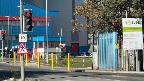 The Suez waste facility is a large blue and grey warehouse, behind metal gates. There is a pedestrian crossing at the entrance and a green and white sign for "suez".