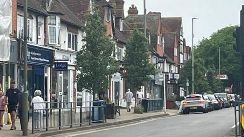 Shops line the road in West Byfleet with railings along pavements, where people are walking past the businesses. There are trees dotted along the road and further in the background. Cars are parked by the roadside and there are lampposts and road signs.
