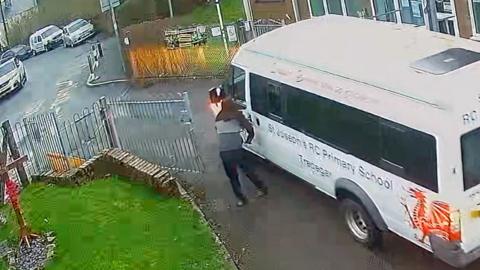 A man with his hood up stands next to the white minibus