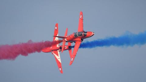 Two red jets pass each other at close distance while releasing red and blue smoke behind them.