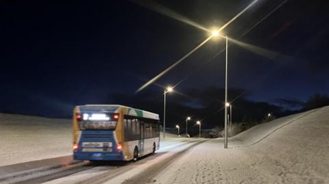 A bus travels along a snow-covered road in Inverness on Wednesday morning. It is still dark and streetlights are on.