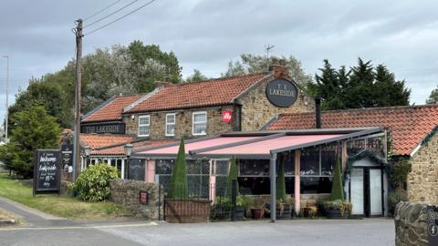 The Lakeside Inn - built in stone with a red-tiled roof, and a pink awning in front of a conservatory. There is a sign for it on the side, and a advertising board near the front entrance