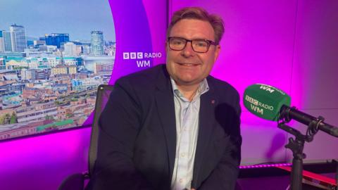 Cllr John Cotton, Labour leader of Birmingham City Council sitting in the BBC WM studio in front of purple studio lighting. He is looking into the camera smiling and leaning against the desk