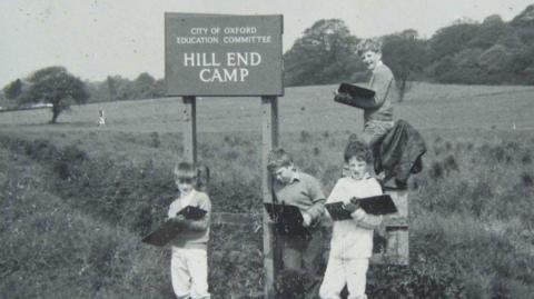 A black and white archive image of school children standing in a field next to a sign for Hill End.