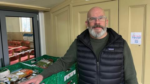Seamus Connolly has a bald head and a full grey beard and moustache, his glasses have clear frames his has a black body warmer over a dark green sweater and is leaning against some green crates of donated foods. Behind him is a room with some chairs.