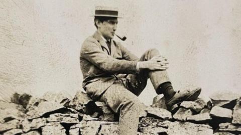Photograph of the Cheshire mountaineer George Mallory. The black and white image shows him sitting on a dry stone wall.