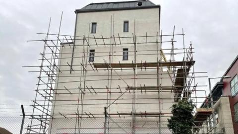 A side profile of the building. It has a white rendering and is covered in scaffolding. There are clouds above.