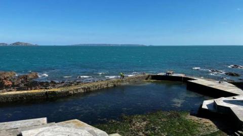 Guernsey Bathing Pools at La Valette. The water is a dark blue. There is seaweed. The sky is light blue. 