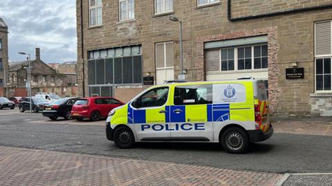 A police van sits in the courtyard of a large building. Other vehicles are parked in the background.